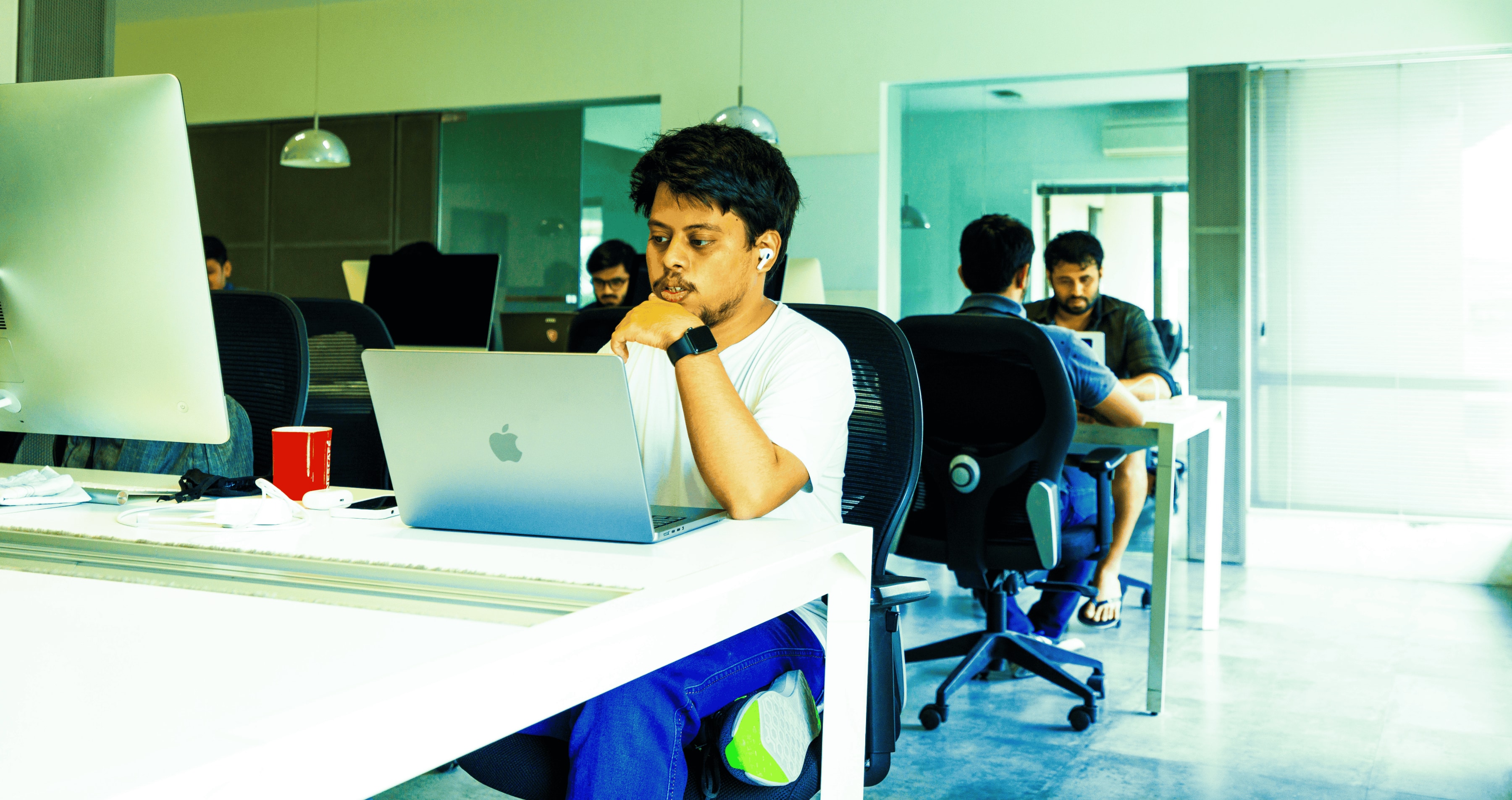 In a brightly lit office, a young man from the design team works diligently on his laptop, surrounded by the focused energy of his teammates, embodying the harmony of design and engineering at Samespace.