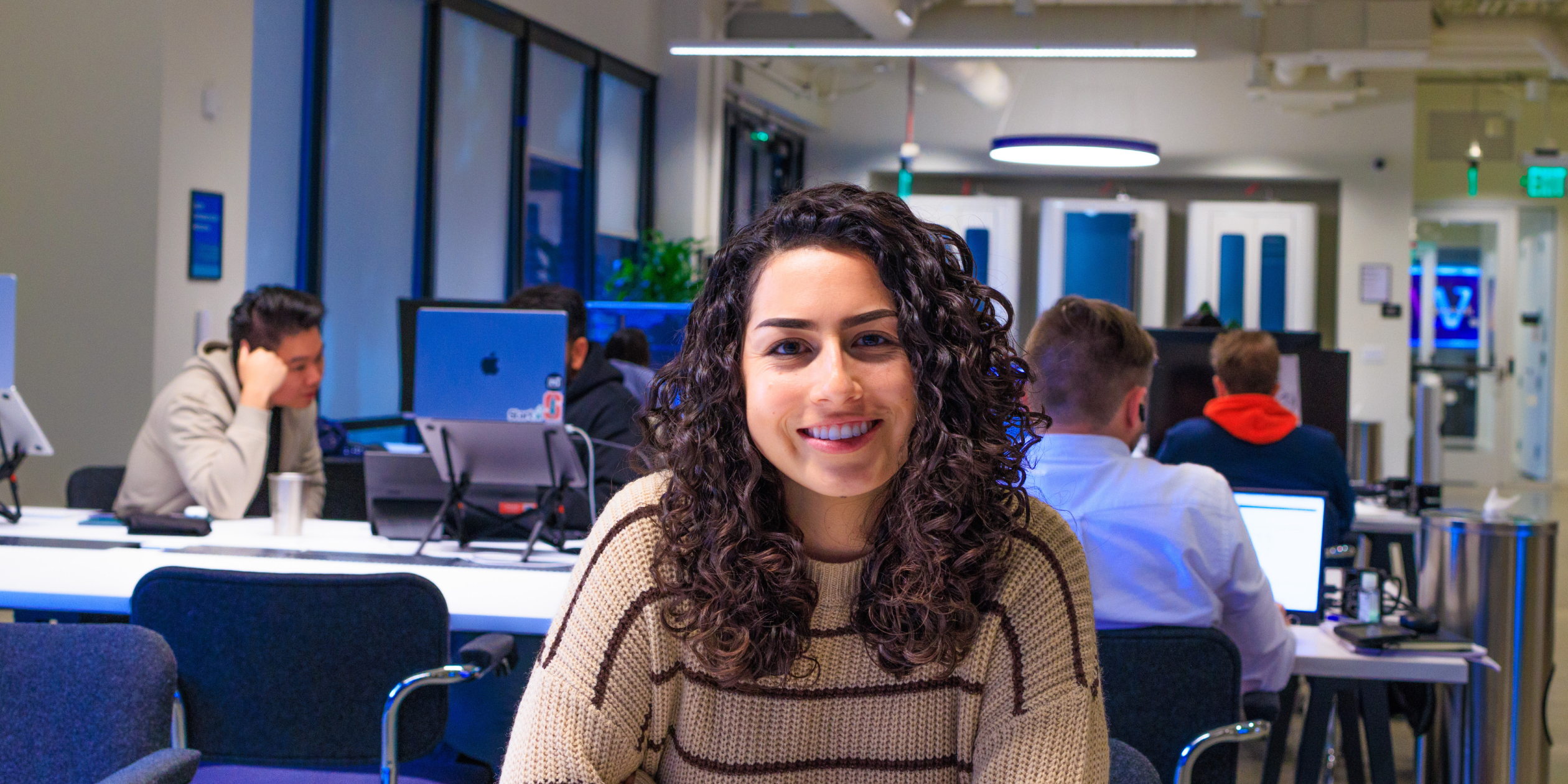 A woman with curly hair smiling at the camera, seated in a vibrant office environment where colleagues can be seen engaged at their workstations, exemplifying Samespace’s dynamic and passionate work culture.